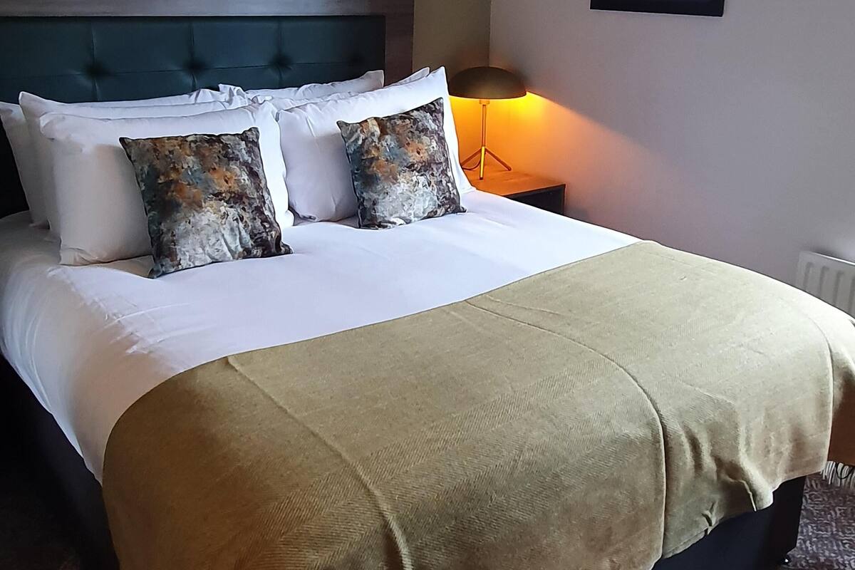 A neatly made bed with a headboard, two pillows, a beige throw blanket, and a bedside lamp.