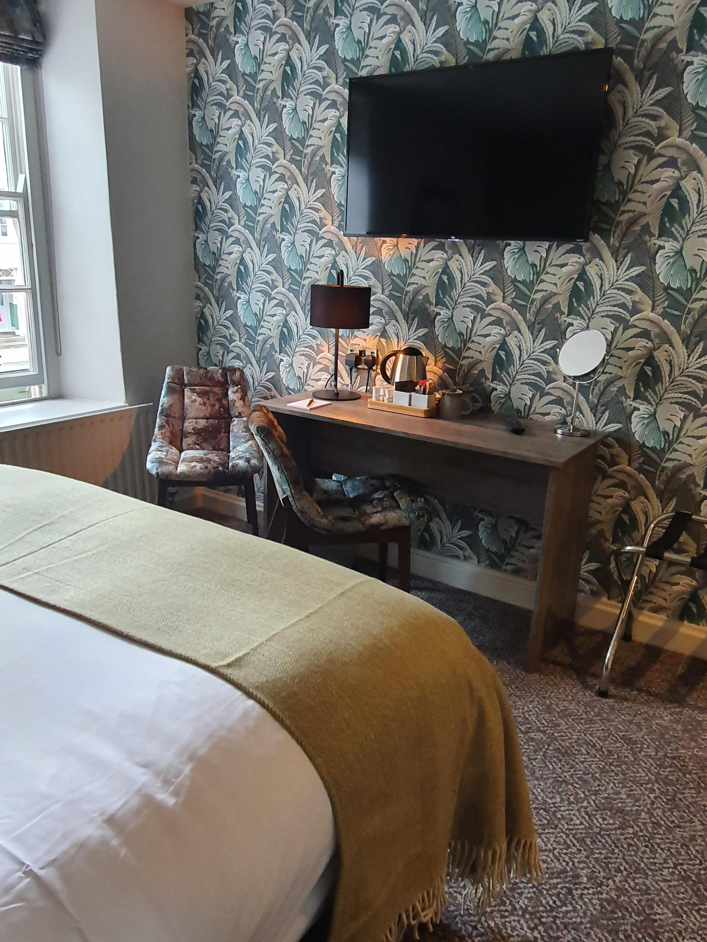 Habitación de hotel con cama, escritorio con lámpara, silla, mesita de noche con espejo y televisor montado en la pared.