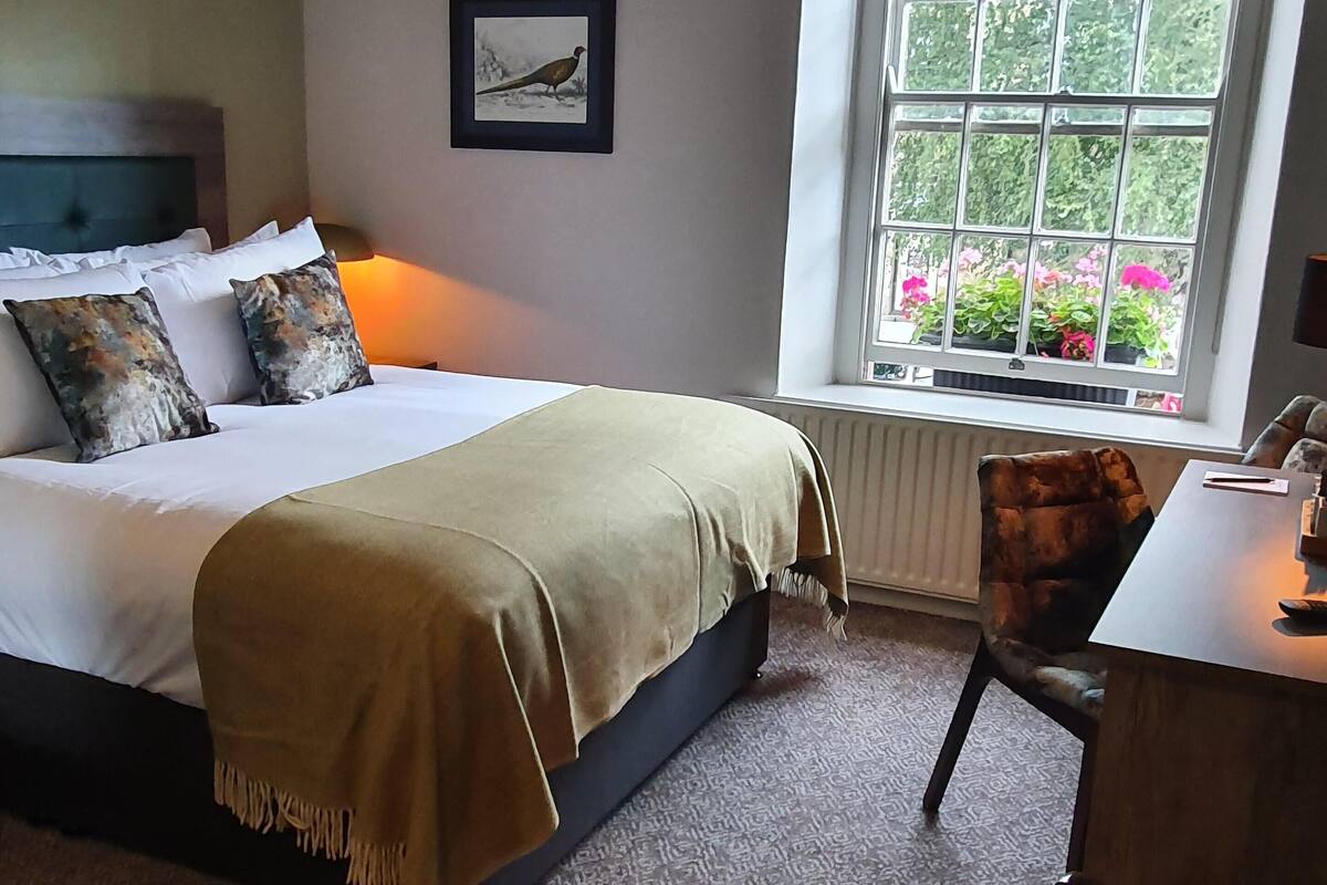 A bedroom with a bed, a desk, a chair, a window with a view of flowers, and a framed picture on the wall.