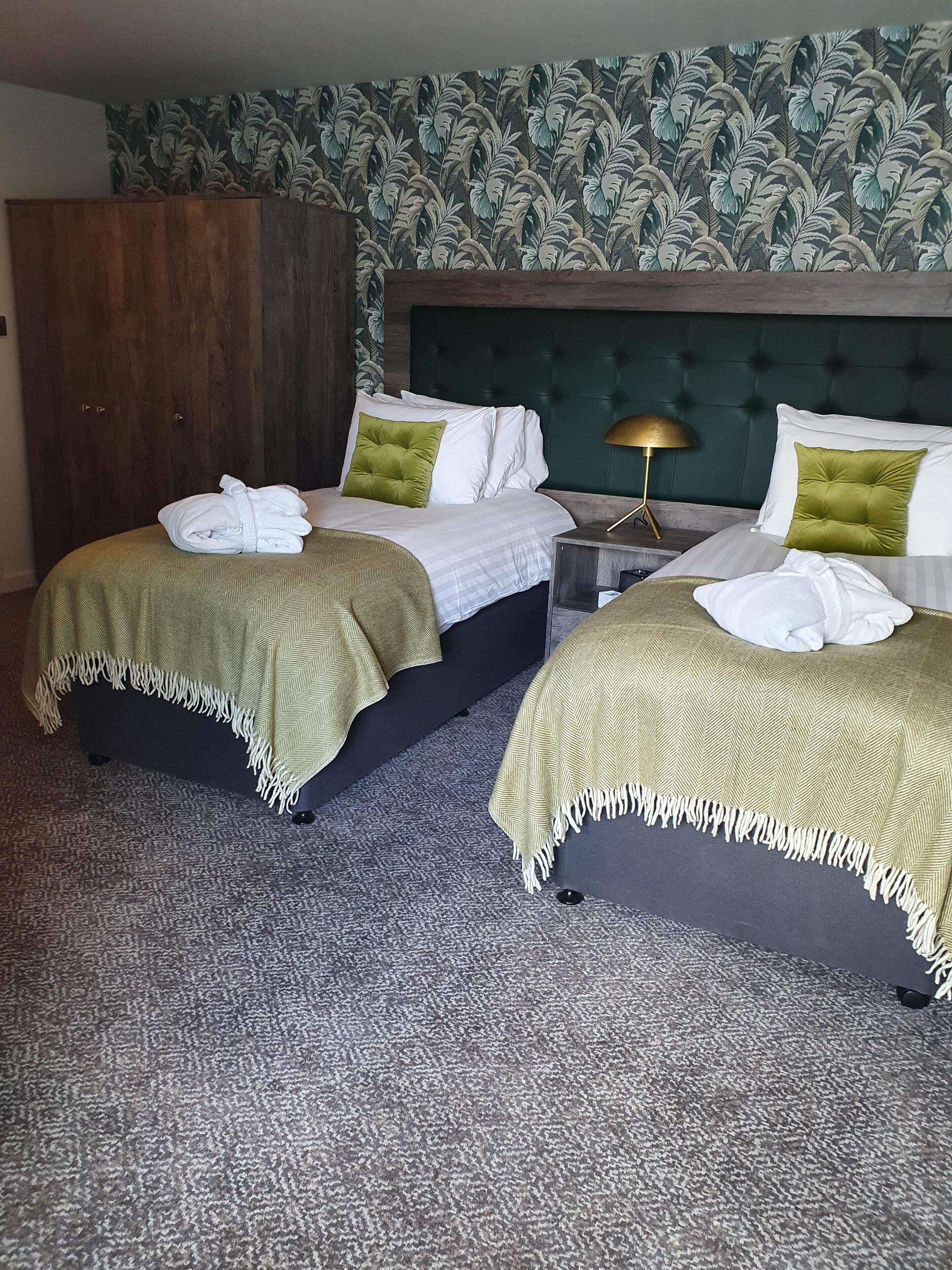 Habitación de hotel con dos camas, cada una con cubrecama verde y almohadas blancas. Se observa un armario de madera y una mesita de noche con lámpara.