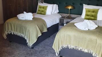 Habitación de hotel con dos camas, cada una con cubrecama verde y almohadas blancas. Se observa un armario de madera y una mesita de noche con lámpara.