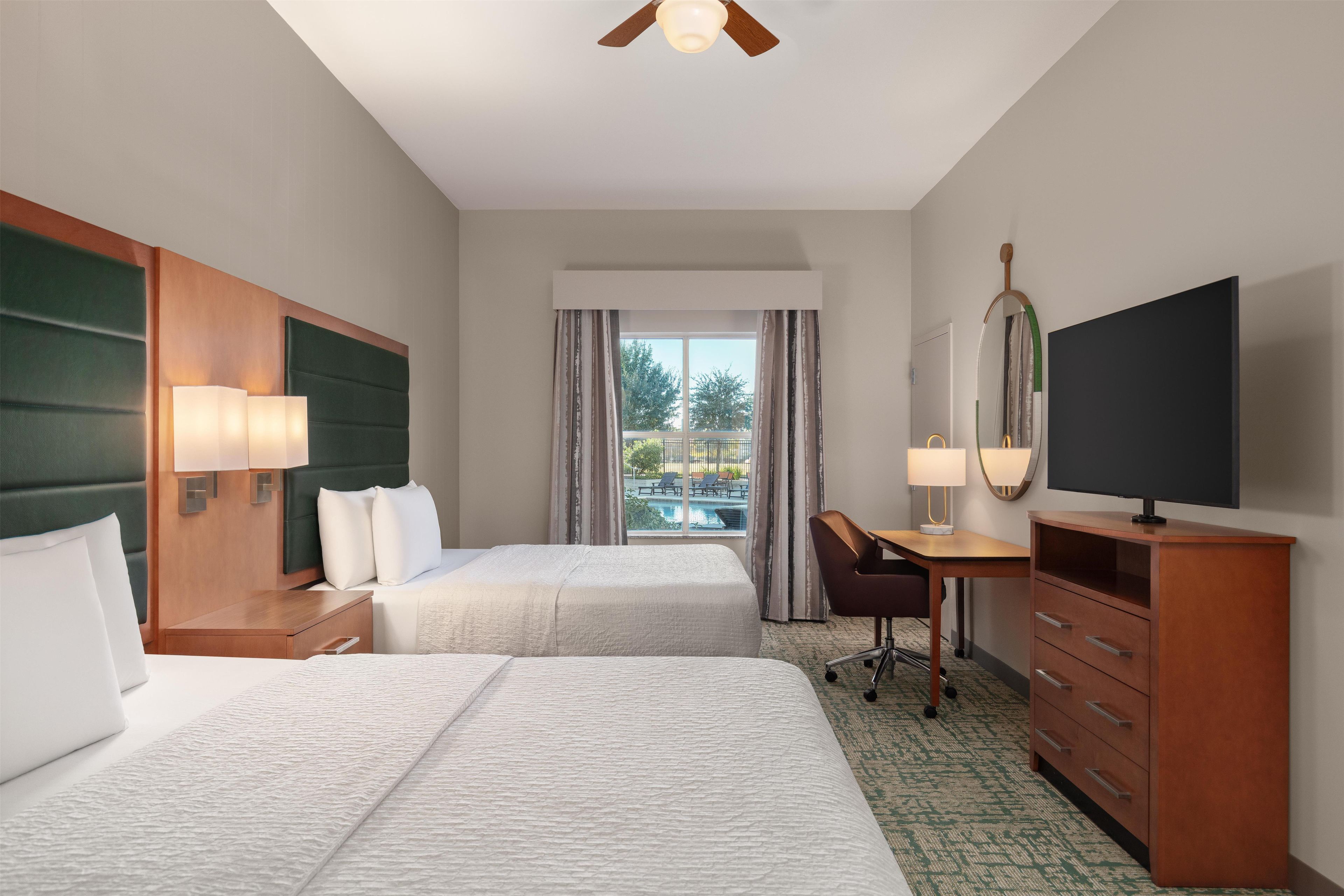 A hotel room with two beds, a desk, a chair, a television, and a view of an outdoor swimming pool.