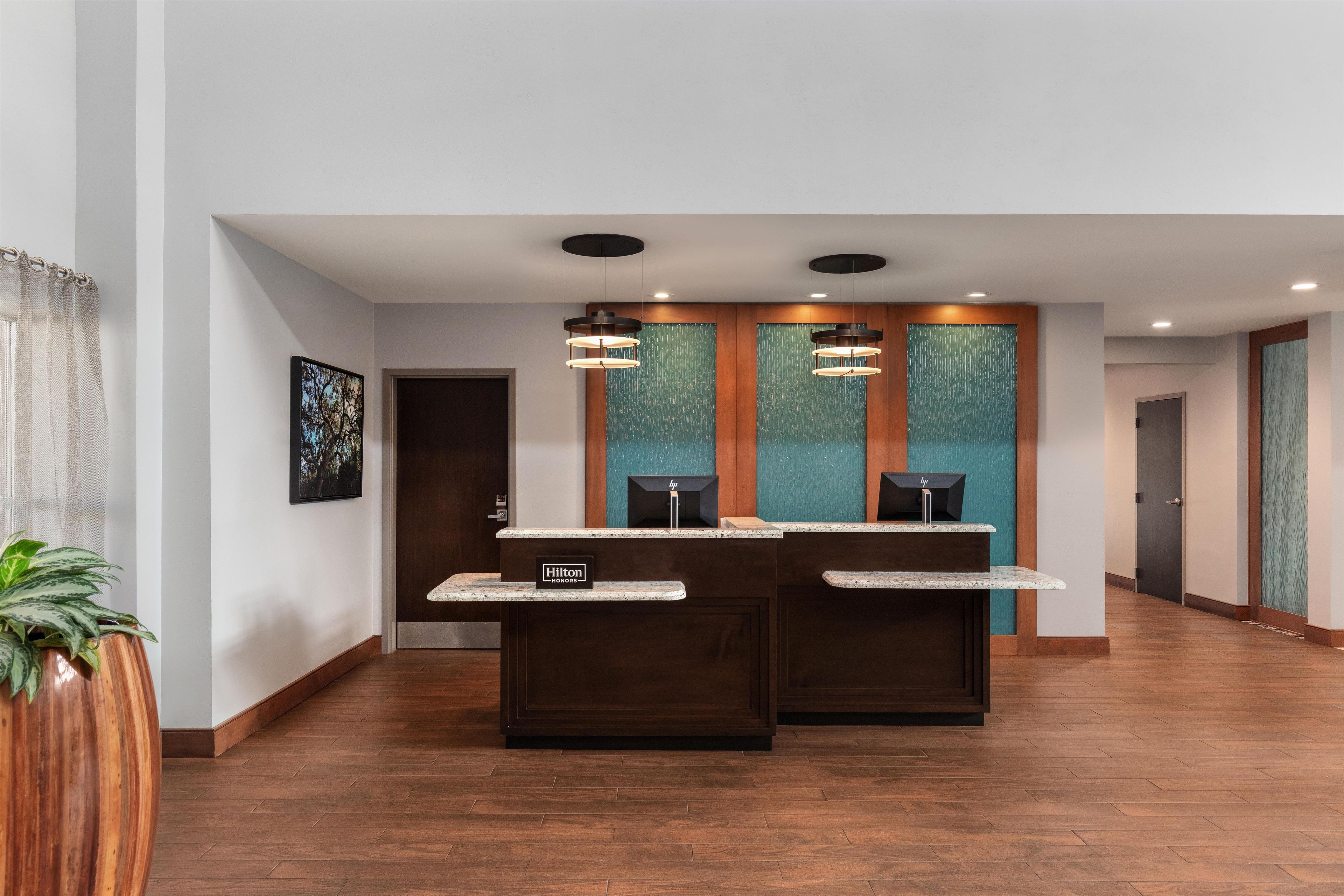 Un espace d’accueil d’hôtel avec un plancher en bois, deux comptoirs de réception, un grand tableau encadré au mur et une plante en pot.