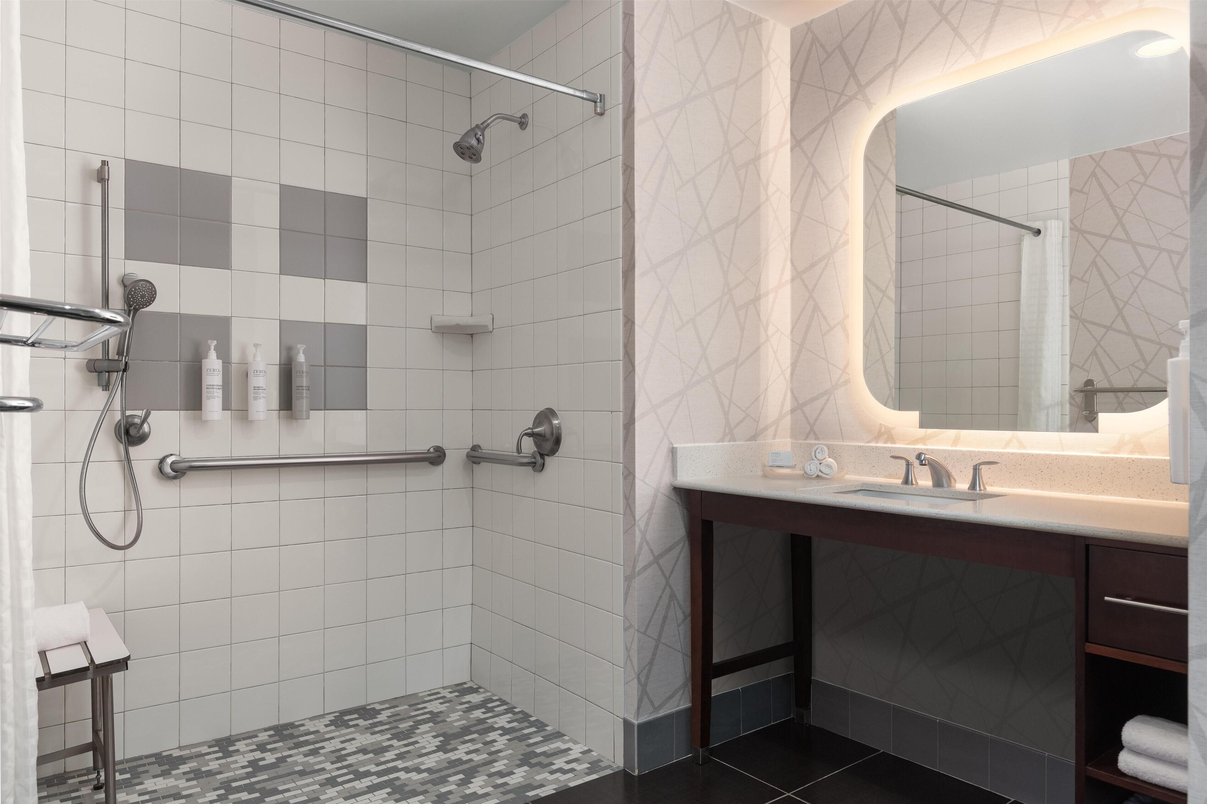 Une salle de bain moderne équipée d’une douche, de barres d’appui, d’un meuble-lavabo avec miroir et d’un sol carrelé.
