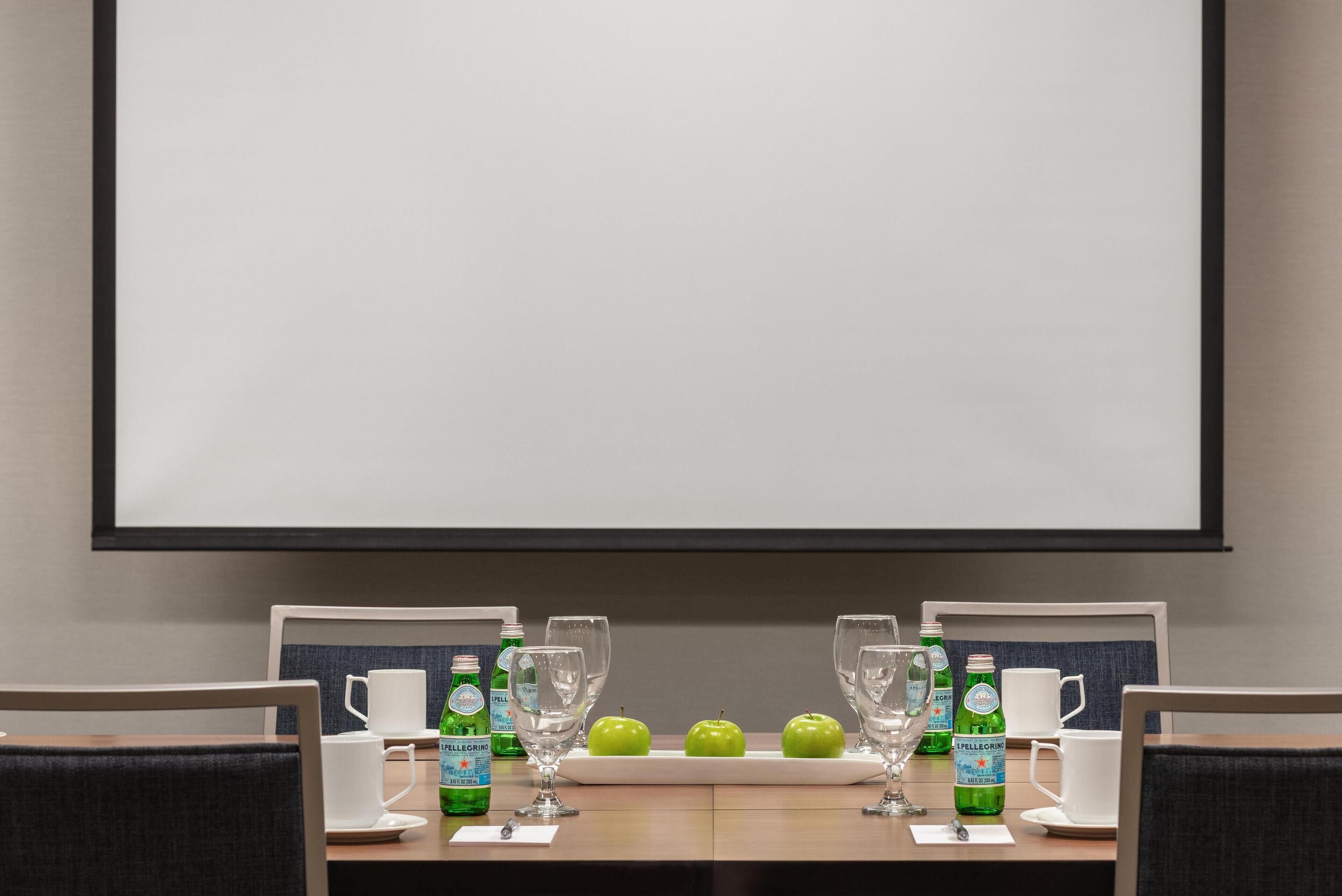A conference room with a large screen, two tables with chairs, and a whiteboard.