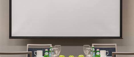 A conference room with a large screen, two tables with chairs, and a whiteboard.