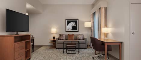 A hotel room with a bed, a TV, a desk, a chair, a sofa, two lamps, and a framed picture on the wall.