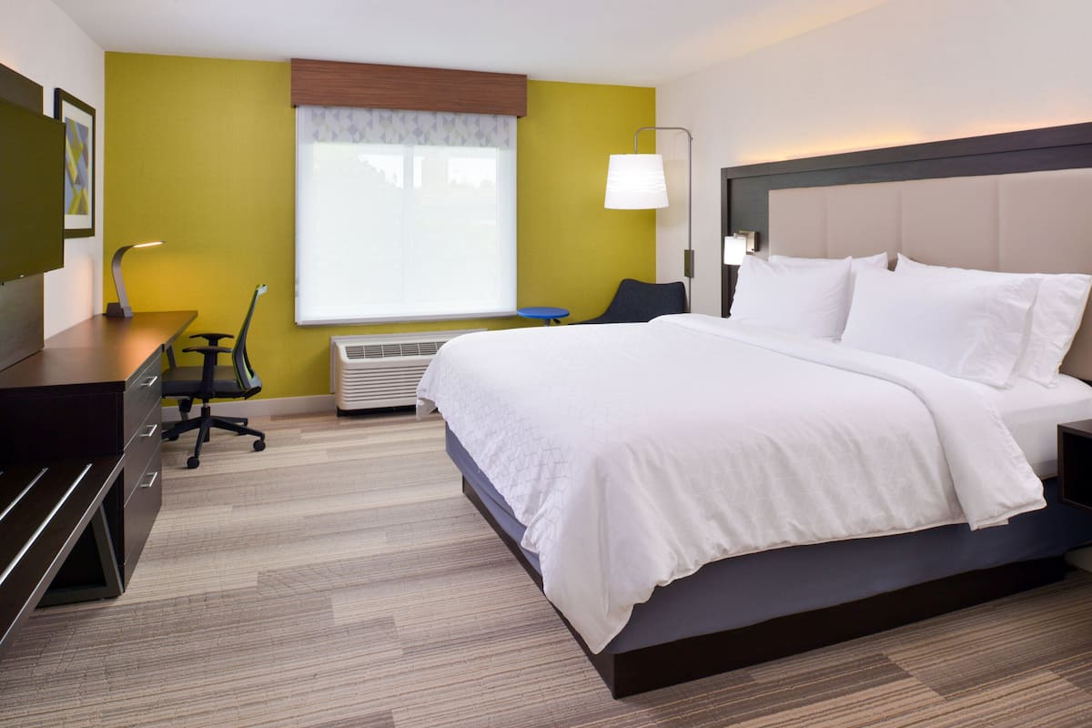Une chambre d’hôtel avec un grand lit, un bureau avec une chaise, une lampe et une fenêtre avec des rideaux.