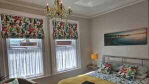 Ein Schlafzimmer mit einem Bett, einem Sessel, einem Nachttisch, einer Lampe, einem Chandelier und zwei Fenstern mit Vorhängen.
