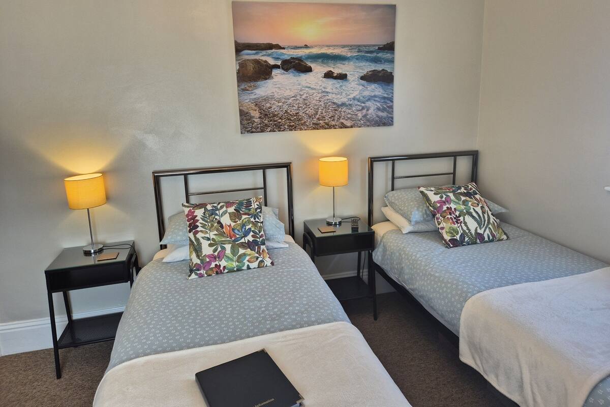 A hotel room with two beds, each with a pillow and a lamp. There is a painting of a sunset on the wall.
