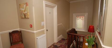 A hallway with patterned carpet, a wooden chair, a table with a vase of flowers, a red lamp, and a door with the number 3.