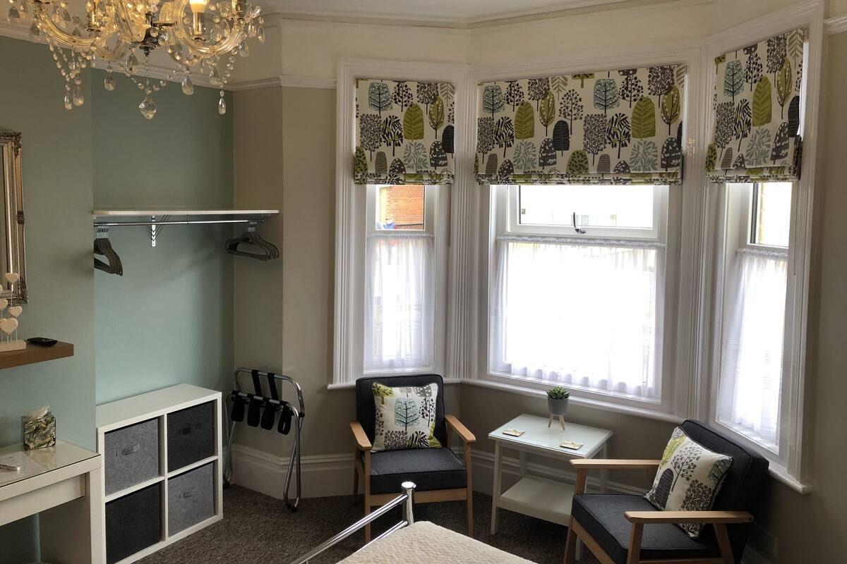 A cozy room with a chandelier, a window with patterned curtains, a chair, a small table, and a coat rack.