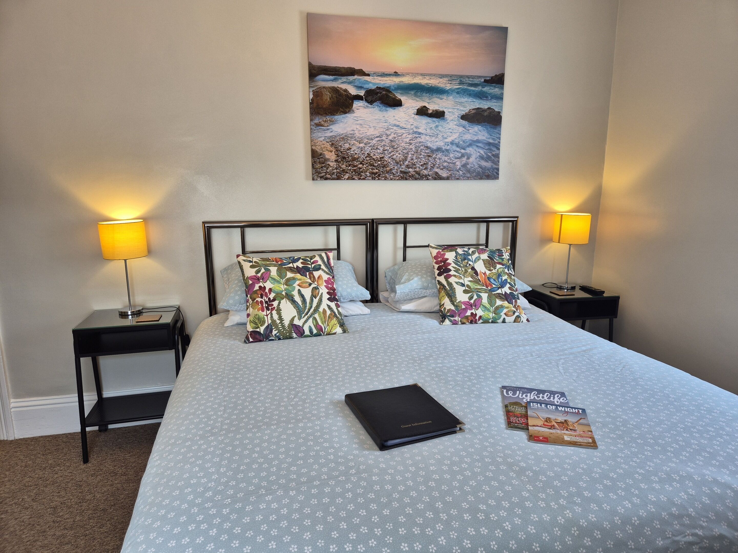 Habitación de hotel con cama, dos mesitas de noche con lámparas, una mesita auxiliar y un cuadro enmarcado con una escena de playa, además de una guía de viaje sobre la cama.