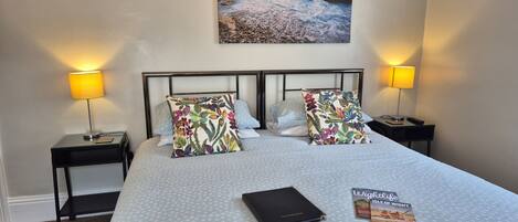 Habitación de hotel con cama, dos mesitas de noche con lámparas, una mesita auxiliar y un cuadro enmarcado con una escena de playa, además de una guía de viaje sobre la cama.