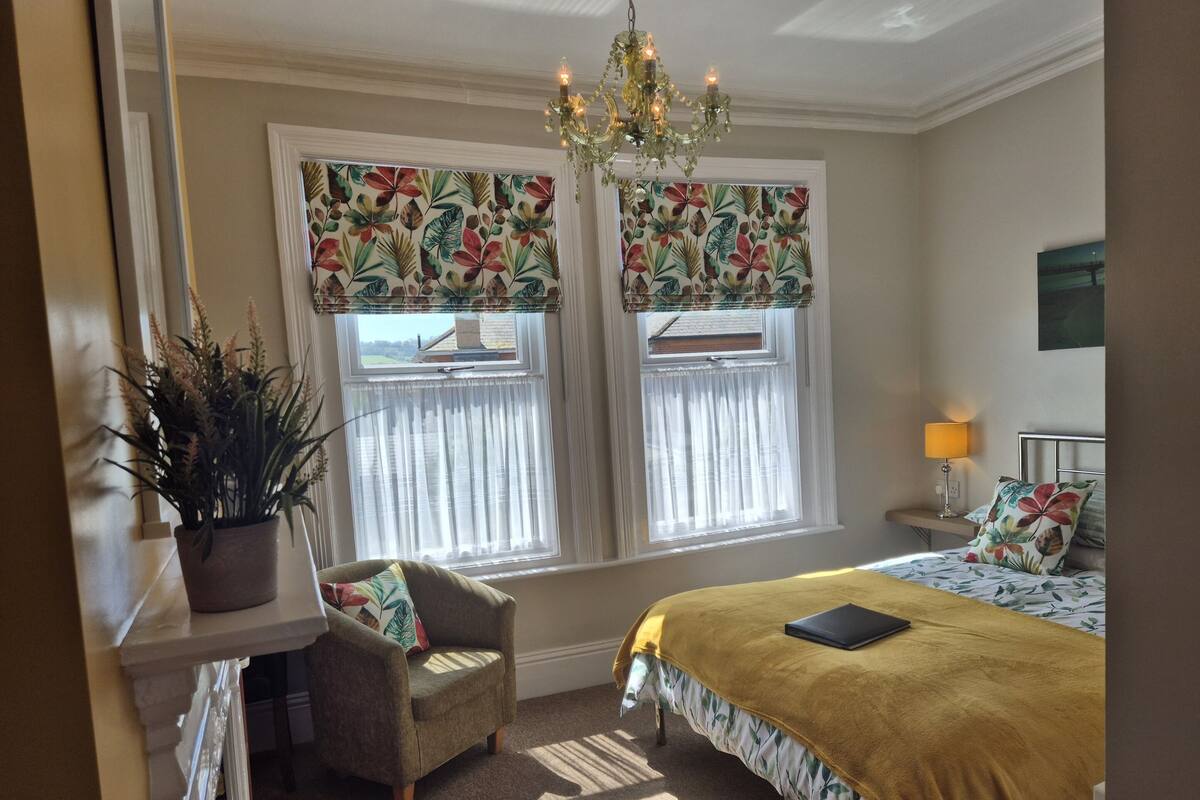 A bedroom with a bed, a chair, a nightstand, a lamp, a potted plant, and two windows with curtains.