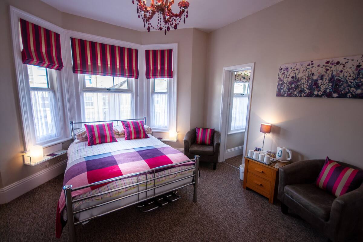 A bedroom with a metal bed frame, a checkered bedspread, striped window valances, a brown armchair, a wooden nightstand, and a floral painting on the wall.