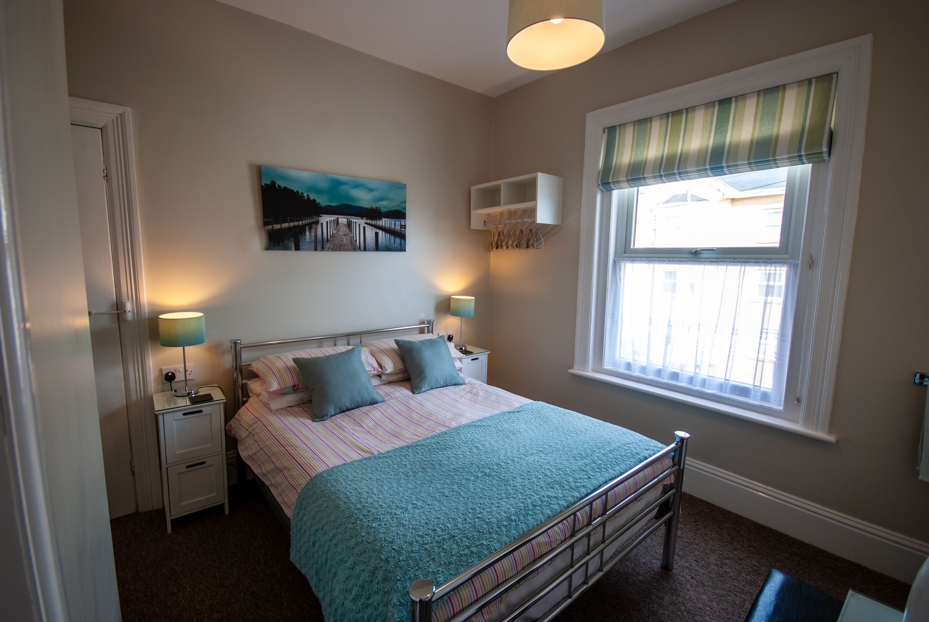 A bedroom with a bed, bedside tables, a lamp, a window with curtains, a painting on the wall, and a view of a building outside.