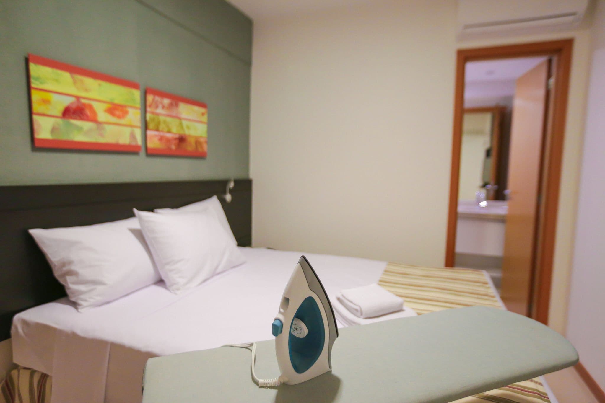 A hotel room with a neatly made bed, an iron, and two colorful paintings on the wall.