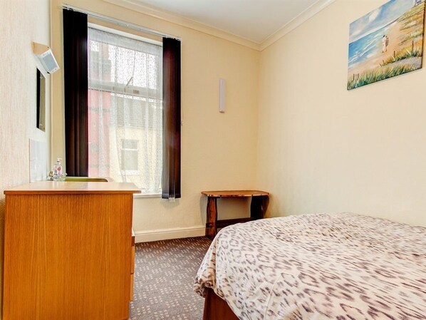 A hotel room with a bed, a wooden desk, a small table, a window with curtains, and a painting on the wall.