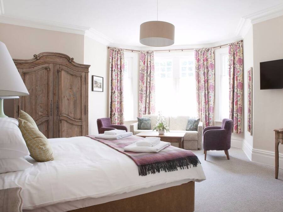 A bedroom with a large bed, a wardrobe, a desk, two chairs, a television, and a window with floral curtains.