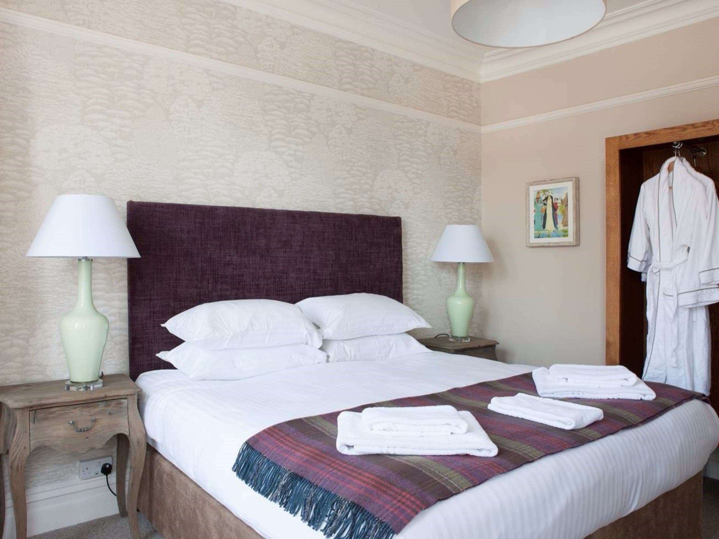 A neatly made bed with white linens, two bedside lamps, a plaid throw blanket, and a white robe hanging on a door.