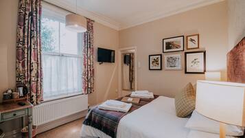 Habitación de hotel con cama, escritorio, radiador, ventana con cortinas, televisor y cuadros en la pared.