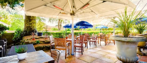 An outdoor patio area with tables and chairs under a large umbrella, surrounded by greenery and potted plants.