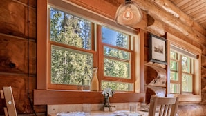A wooden cabin interior with a dining table, chairs, and large windows offering a view of the forest. - Moberly Lodge (Golden)
