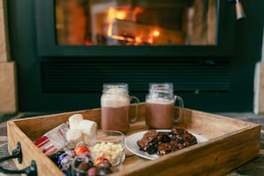 A wooden tray with snacks and drinks, including marshmallows, a glass of milk, and two mugs of hot chocolate, in front of a fireplace.