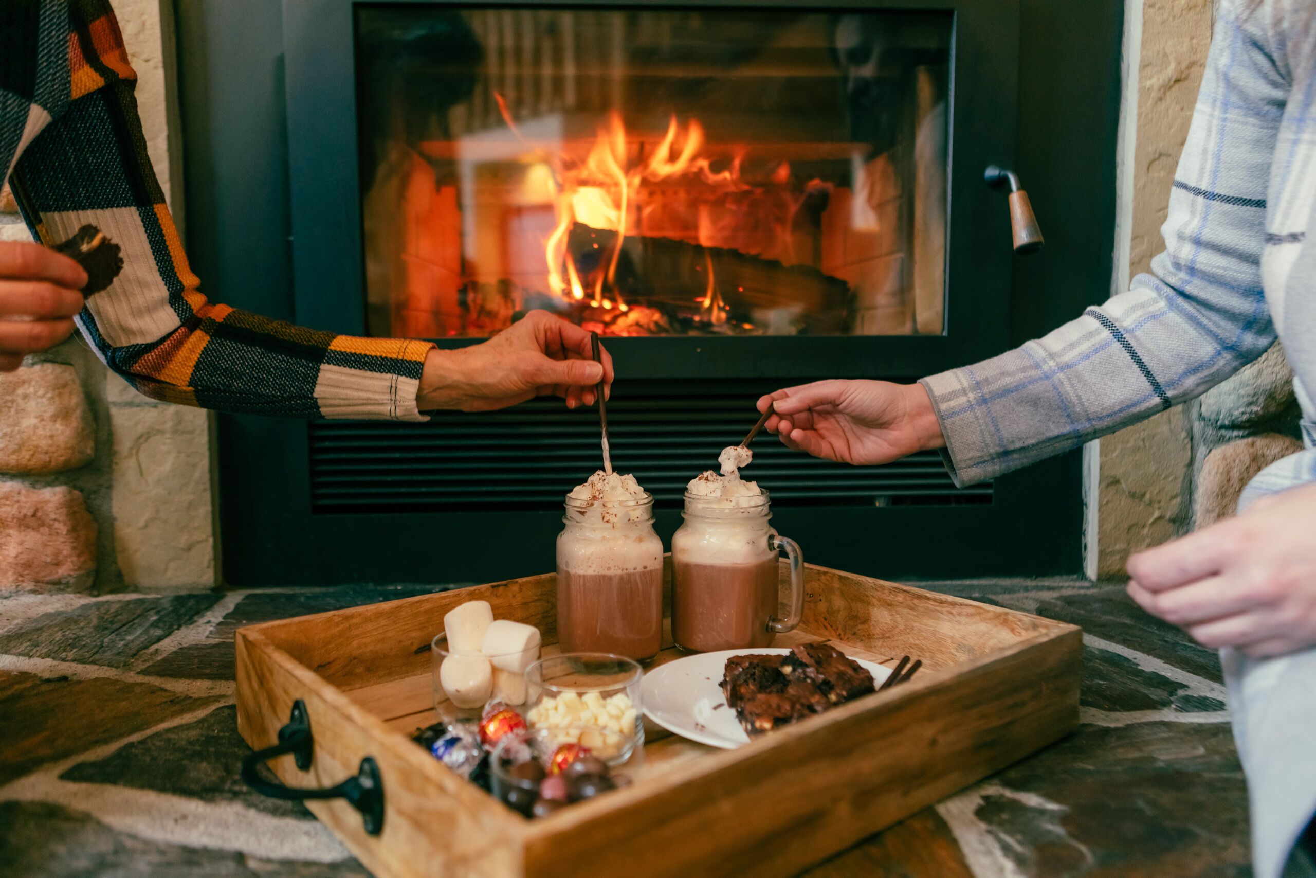 A wooden tray with drinks, snacks, and a plate of food in front of a fireplace.