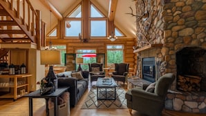 A spacious living room with a stone fireplace, wooden furniture, and large windows offering a view of the outdoors. - Moberly Lodge (Golden)