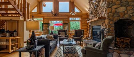 A spacious living room with a stone fireplace, wooden furniture, and large windows offering a view of the outdoors.