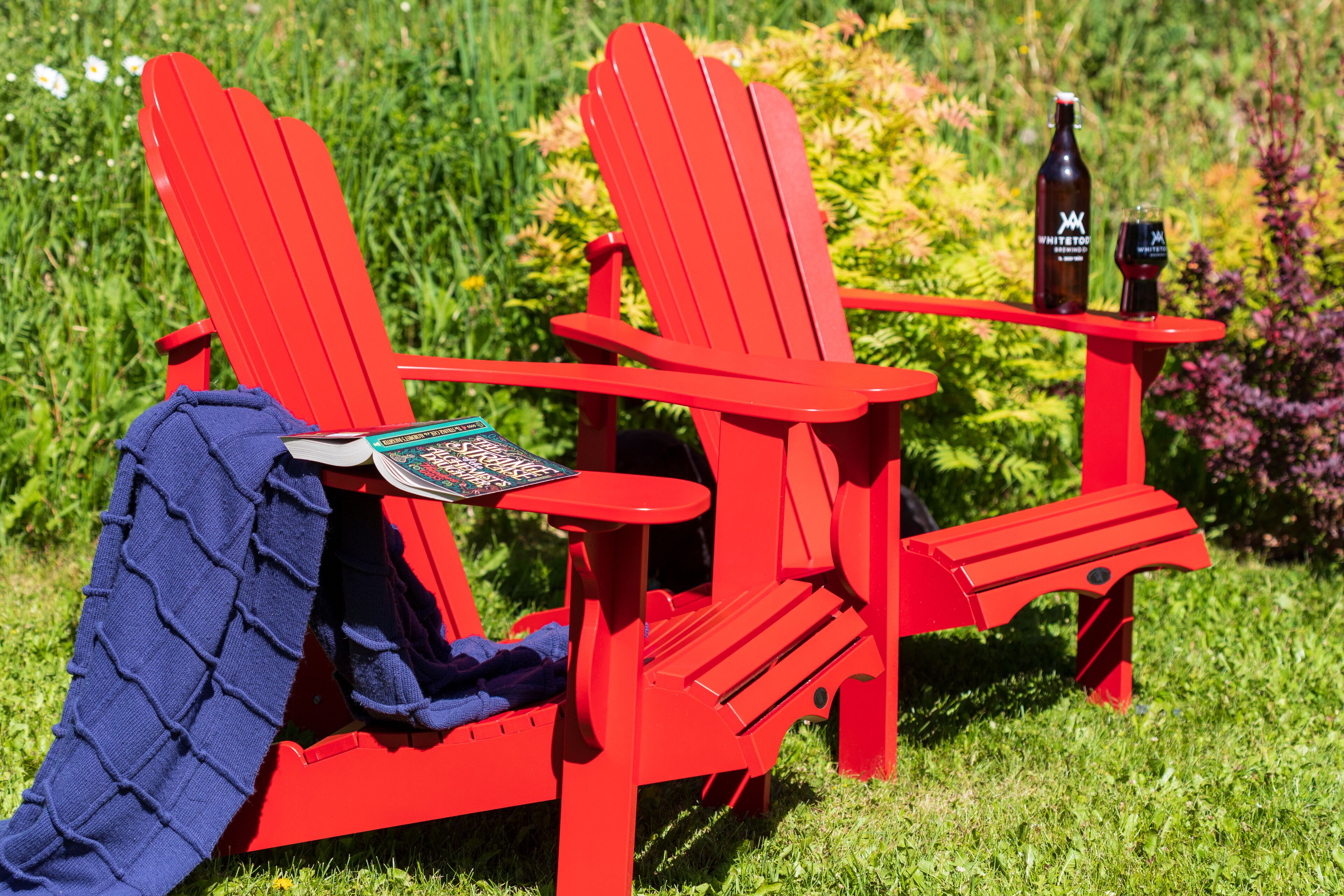 A red Adirondack chair with a blanket and a book, placed on a grassy area with a bottle and glass on a small table beside it.