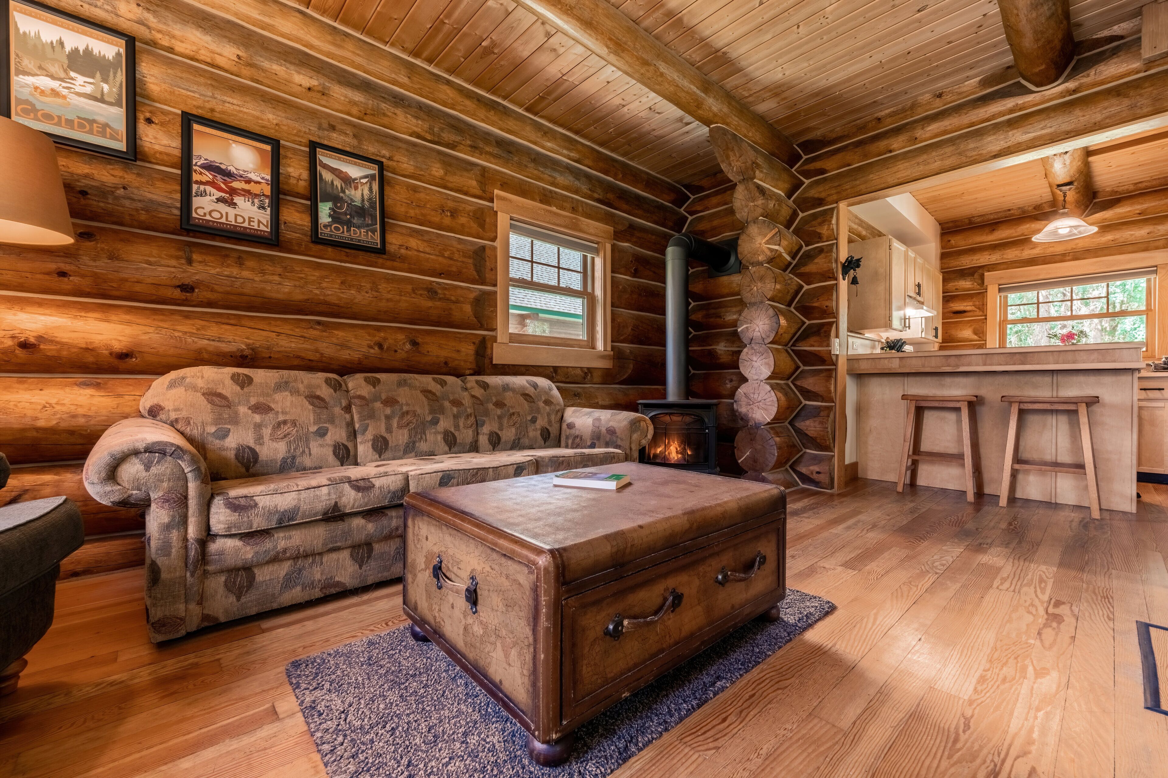 A log cabin interior with a wooden sofa, a chest of drawers, and a kitchen area with stools.