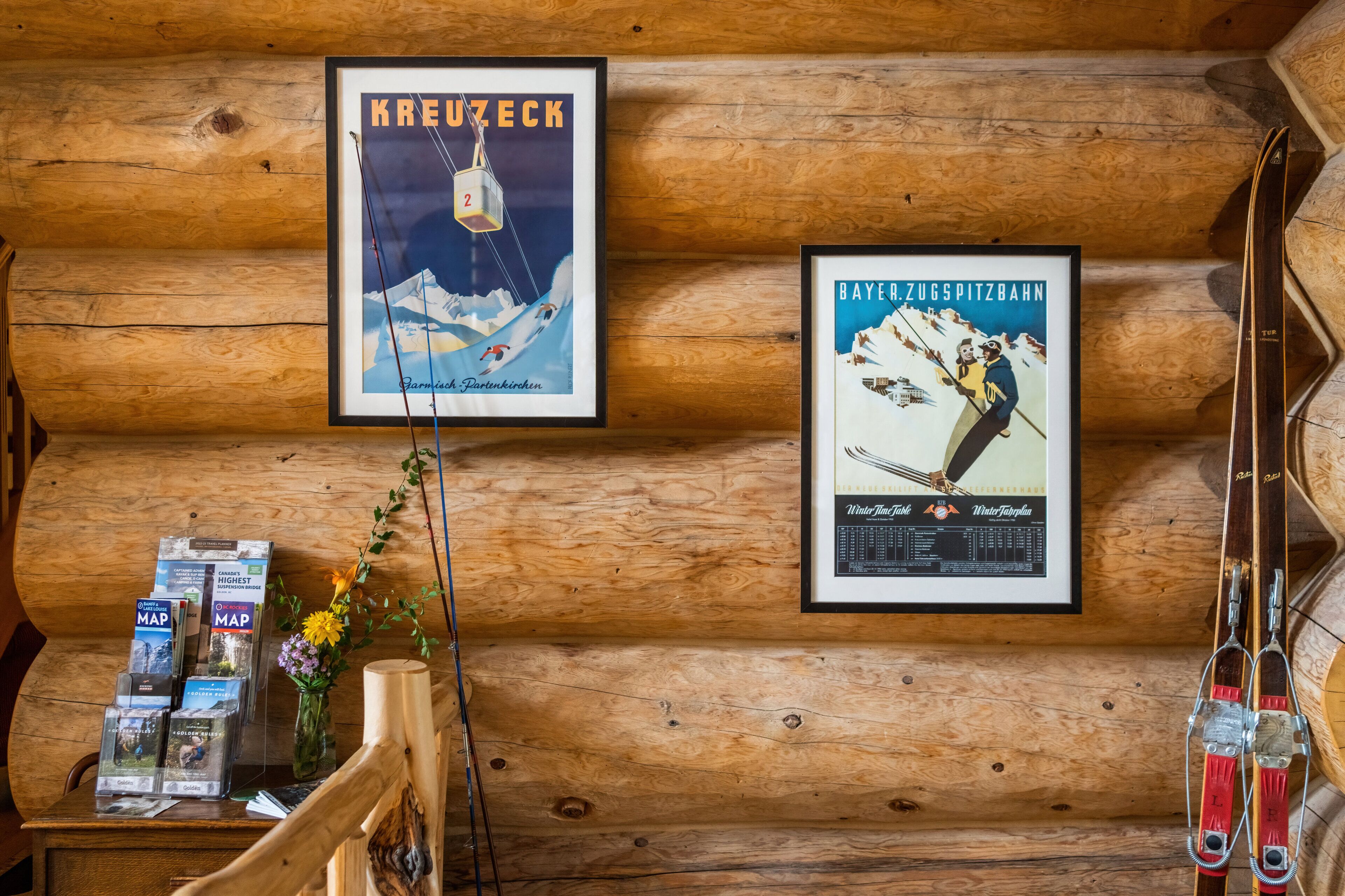 A wooden cabin interior with framed posters of ski destinations, a wooden bench, and a table with ski maps and a vase with flowers.