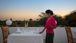 A dining table set for two with white tablecloth, plates, cutlery, and a vase with flowers. A person is standing beside the table, holding a tray.