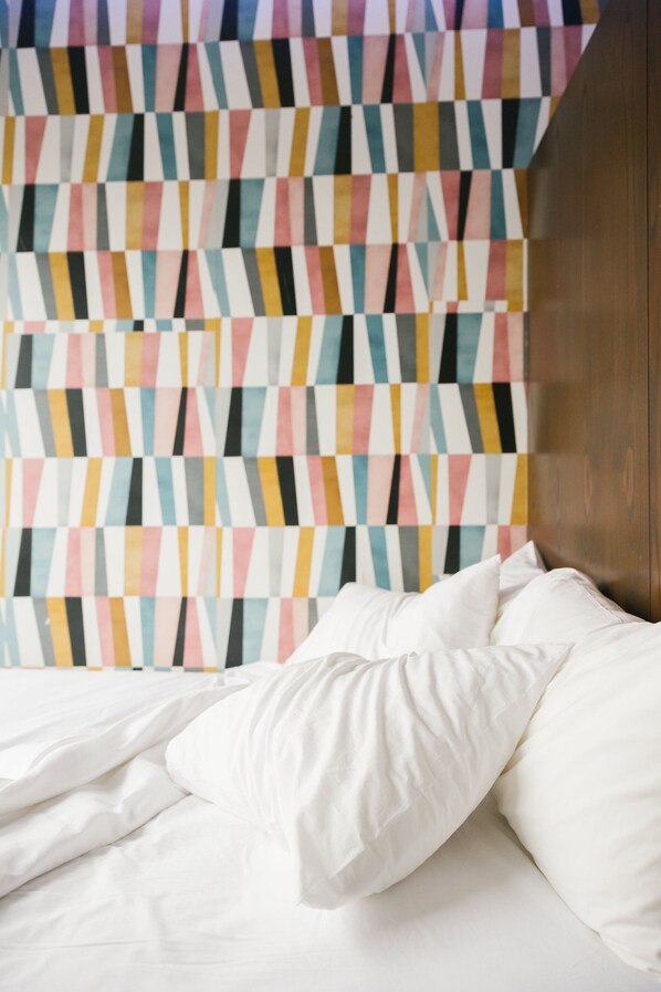 A bed with white bedding and pillows, a colorful geometric-patterned wall behind it, and a wooden headboard. - Orchard Street Hotel (New York)