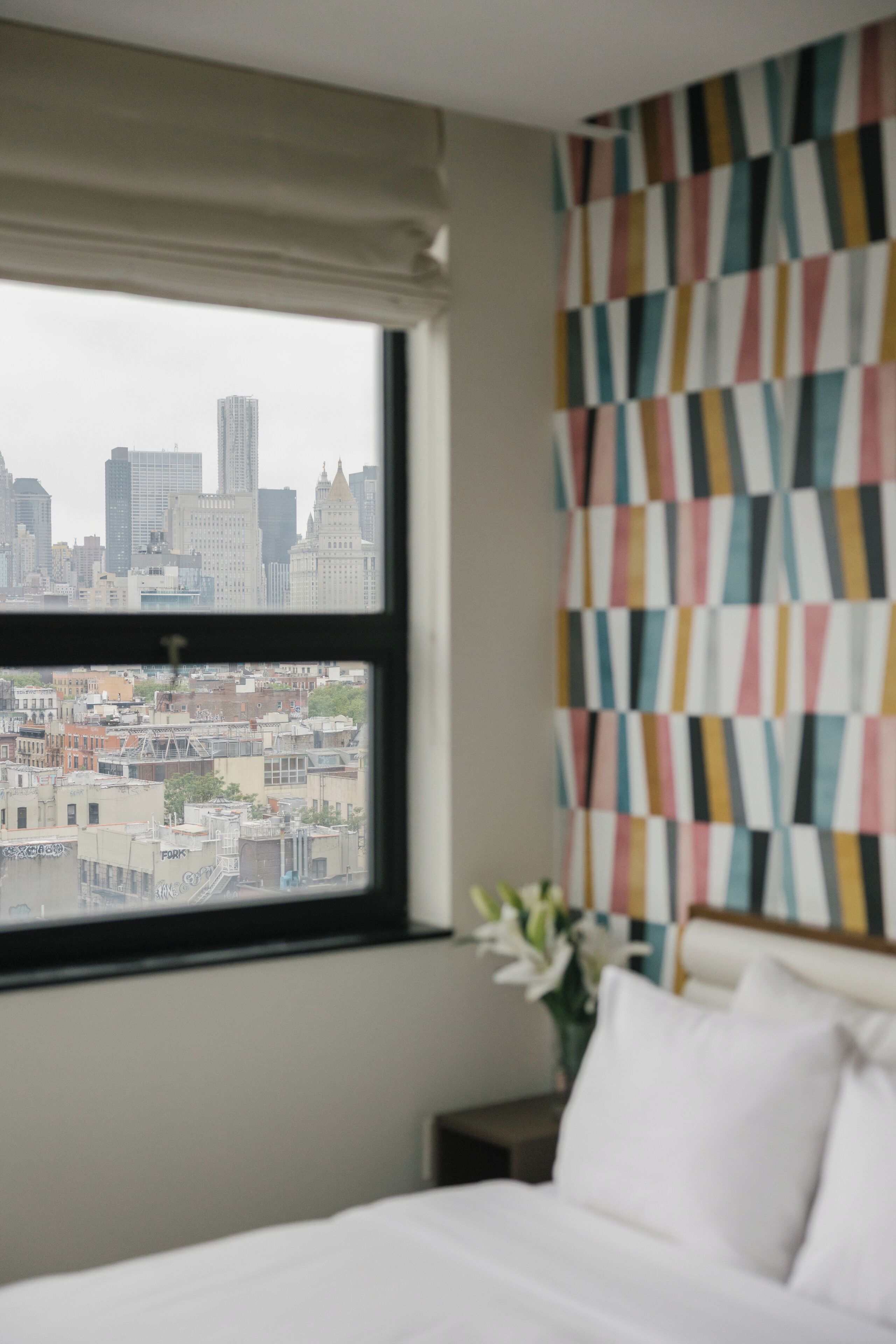 Habitación de hotel con un ventanal amplio que ofrece vistas a la ciudad, una cama, una mesita de noche y una pared con un motivo colorido.