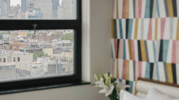 Habitación de hotel con un ventanal amplio que ofrece vistas a la ciudad, una cama, una mesita de noche y una pared con un motivo colorido.