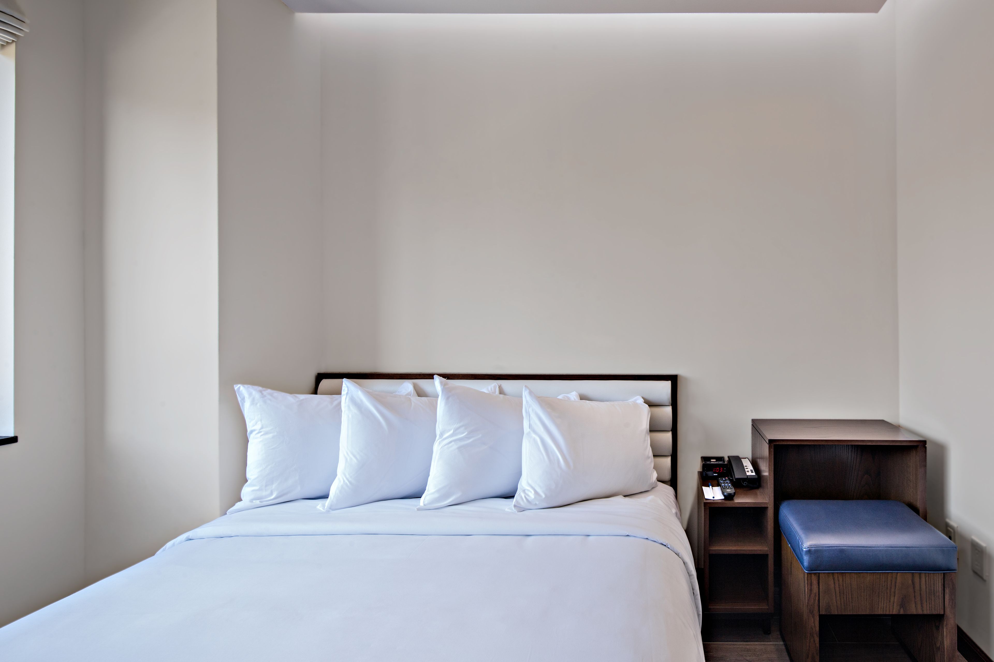 Una cama bien hecha con ropa blanca y almohadas, una mesita de noche de madera con un banco azul, y un teléfono de pared.