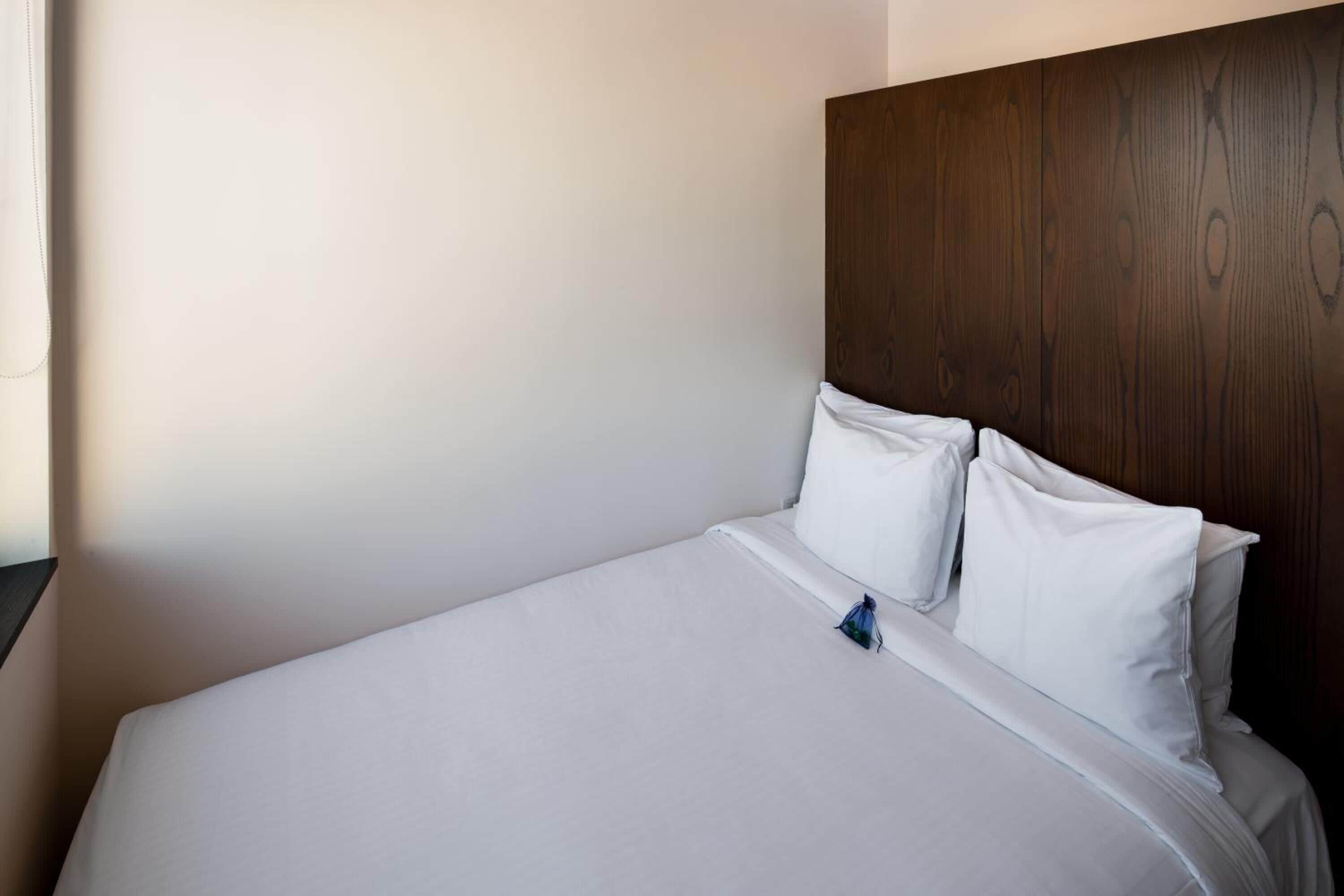 Cama bem arrumada com lençóis e almofadas brancas, cabeceira de madeira e parede lisa.