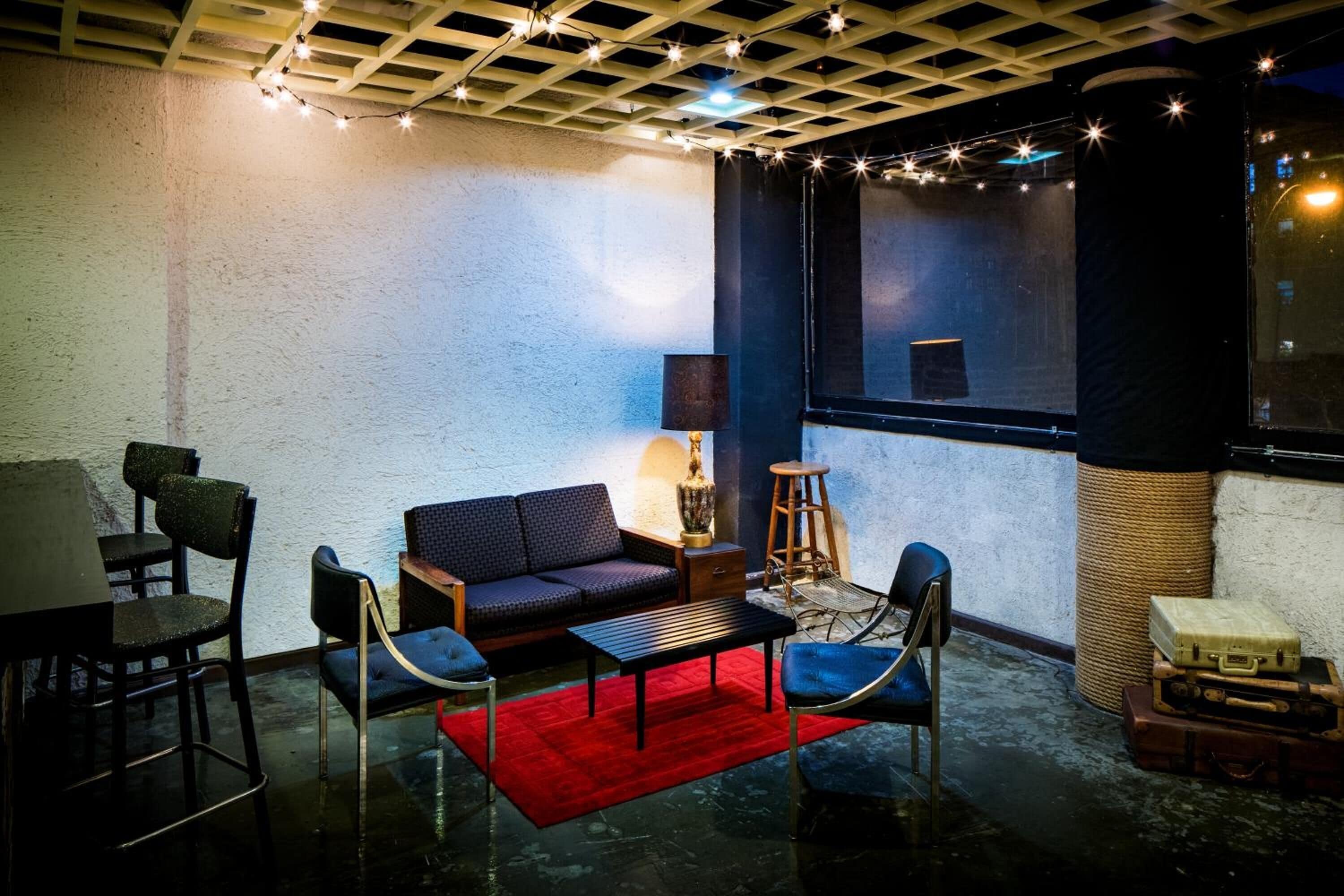 A cozy room with a sofa, chairs, a small table, and a red rug. There is a lamp, a window with curtains, and a ceiling with string lights.