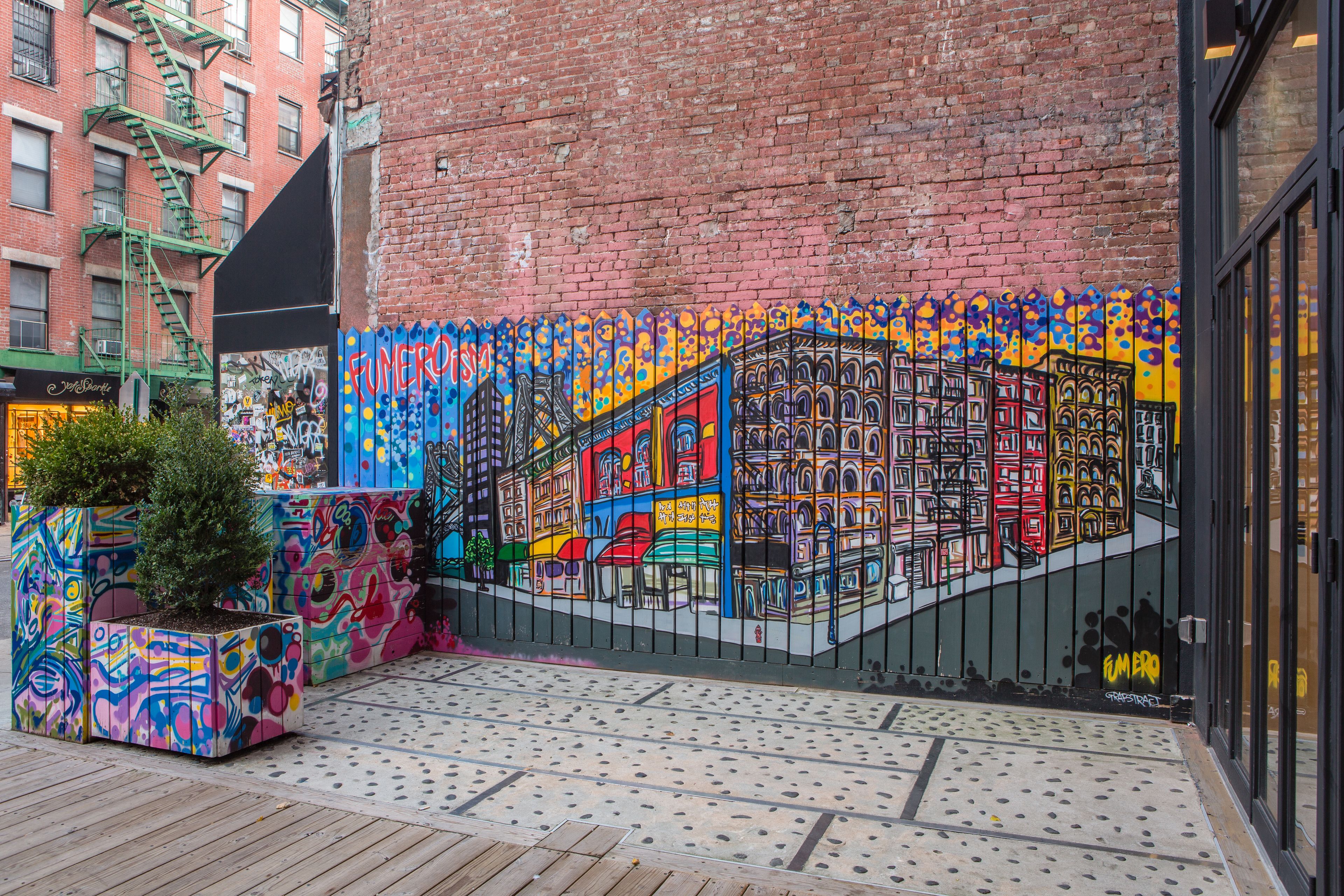 Une fresque colorée orne un bâtiment en briques, on aperçoit une échelle de secours et un jardinier avec un petit arbre.
