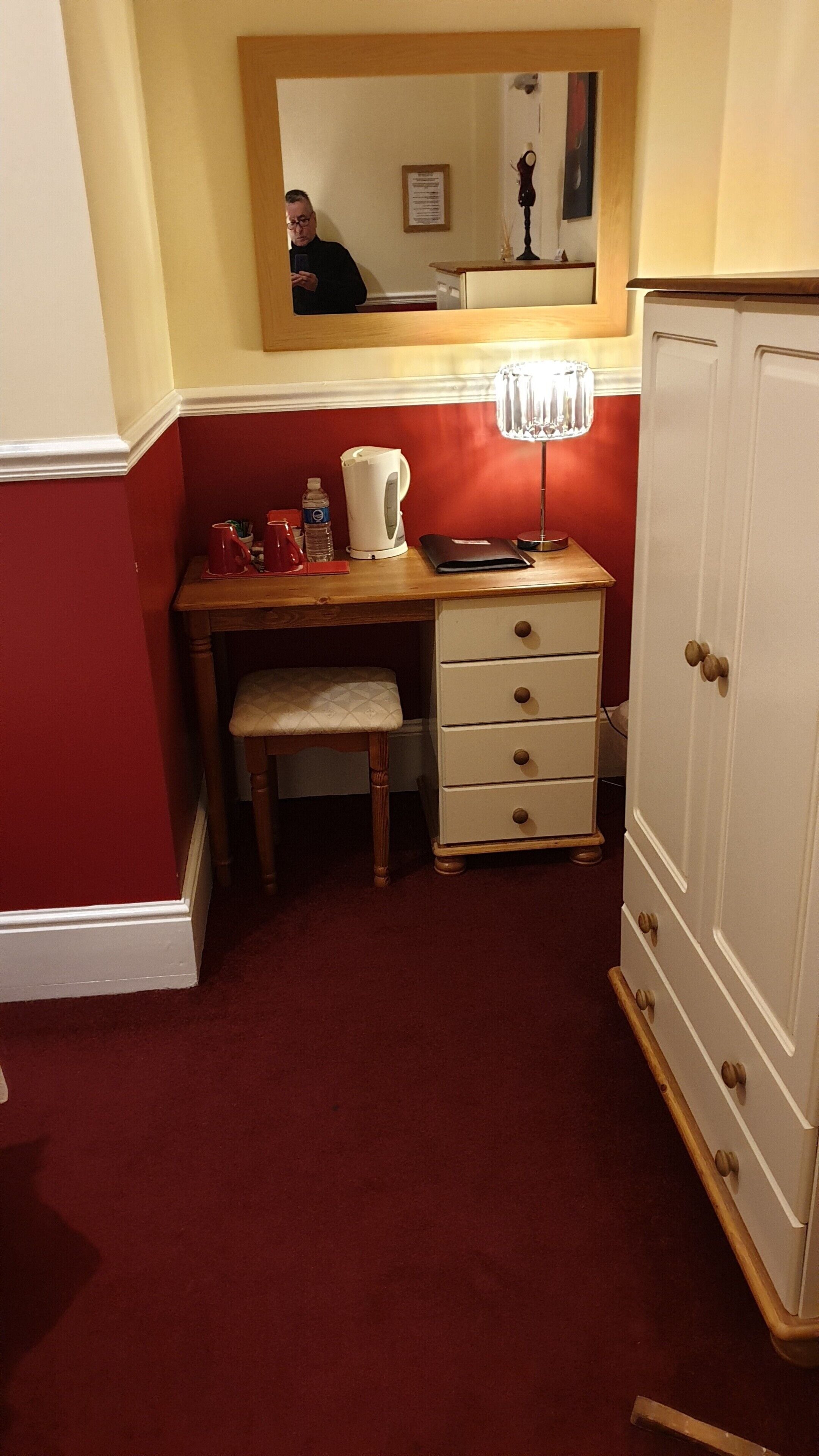 A small room with a wooden desk, a chair, a mirror, a cabinet, and a red carpet.
