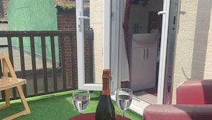 A table with a bottle and two glasses on a red tablecloth, surrounded by red chairs, with a green artificial grass floor and a wooden chair in the background. - All Seasons Guest House (Great Yarmouth)