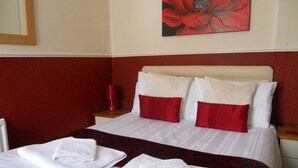 A neatly made bed with white linens and red pillows, a red flower painting on the wall, and a wooden nightstand with a lamp. - All Seasons Guest House (Great Yarmouth)