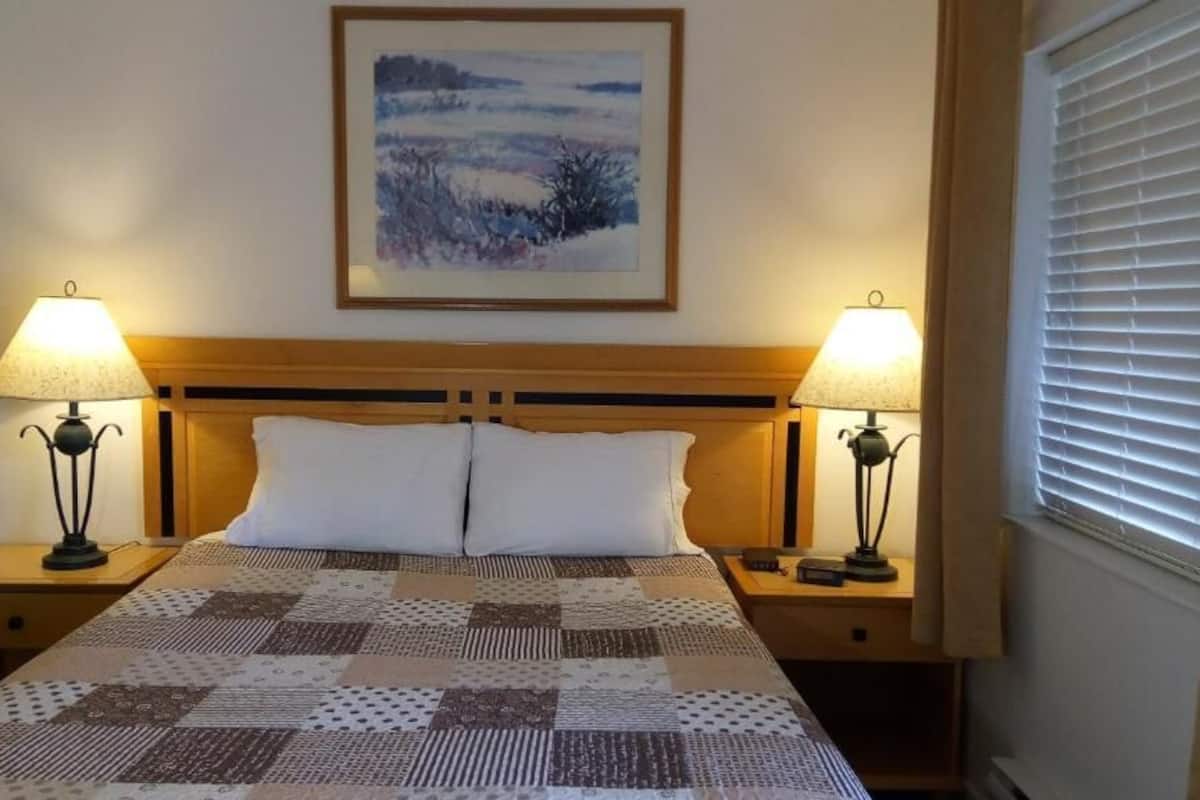 Una cama bien tendida con colcha, dos lámparas de noche, un cuadro enmarcado sobre la cama y una ventana con persianas.