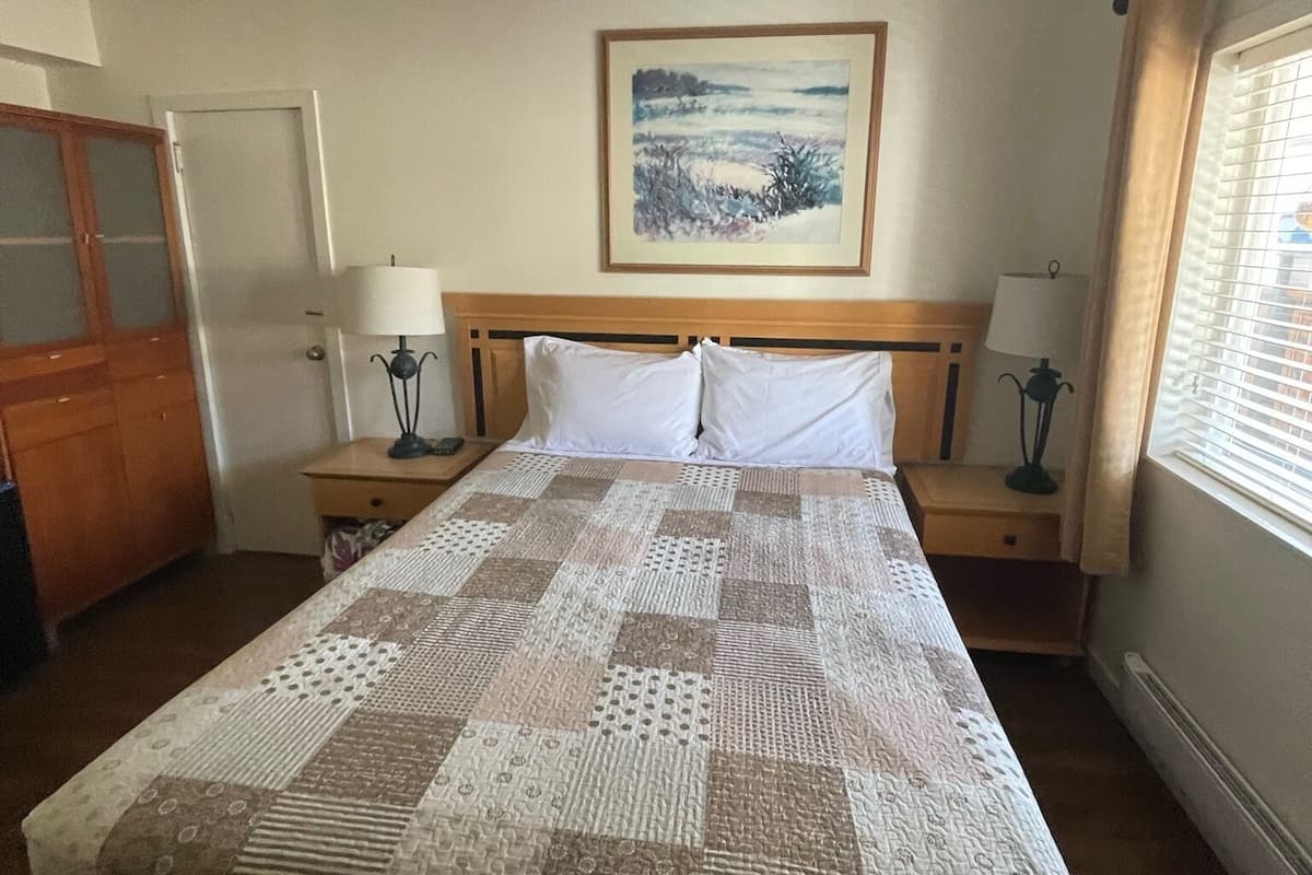 Una habitación de hotel con una cama grande, dos mesitas de noche con lámparas, un cabecero de madera, un cuadro en la pared y una ventana con cortinas.