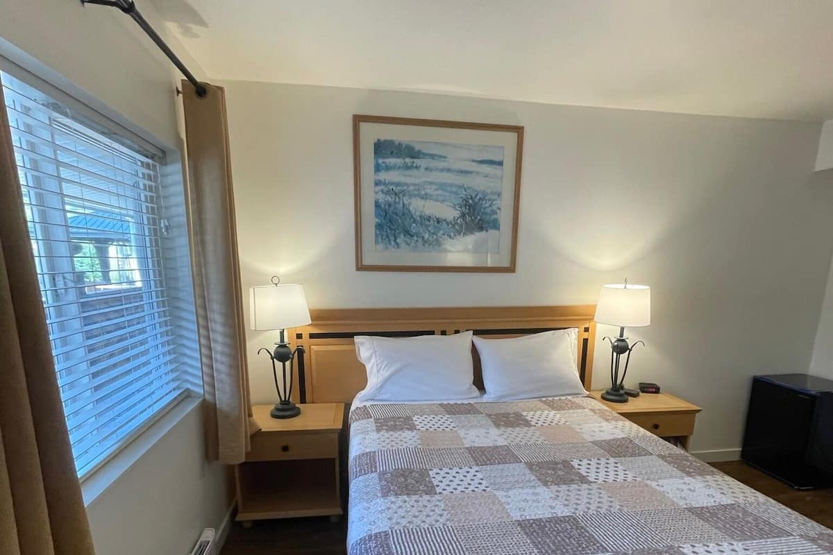 Habitación de hotel con una cama grande, dos mesitas de noche, una lámpara, un cuadro en la pared y una ventana con cortinas.