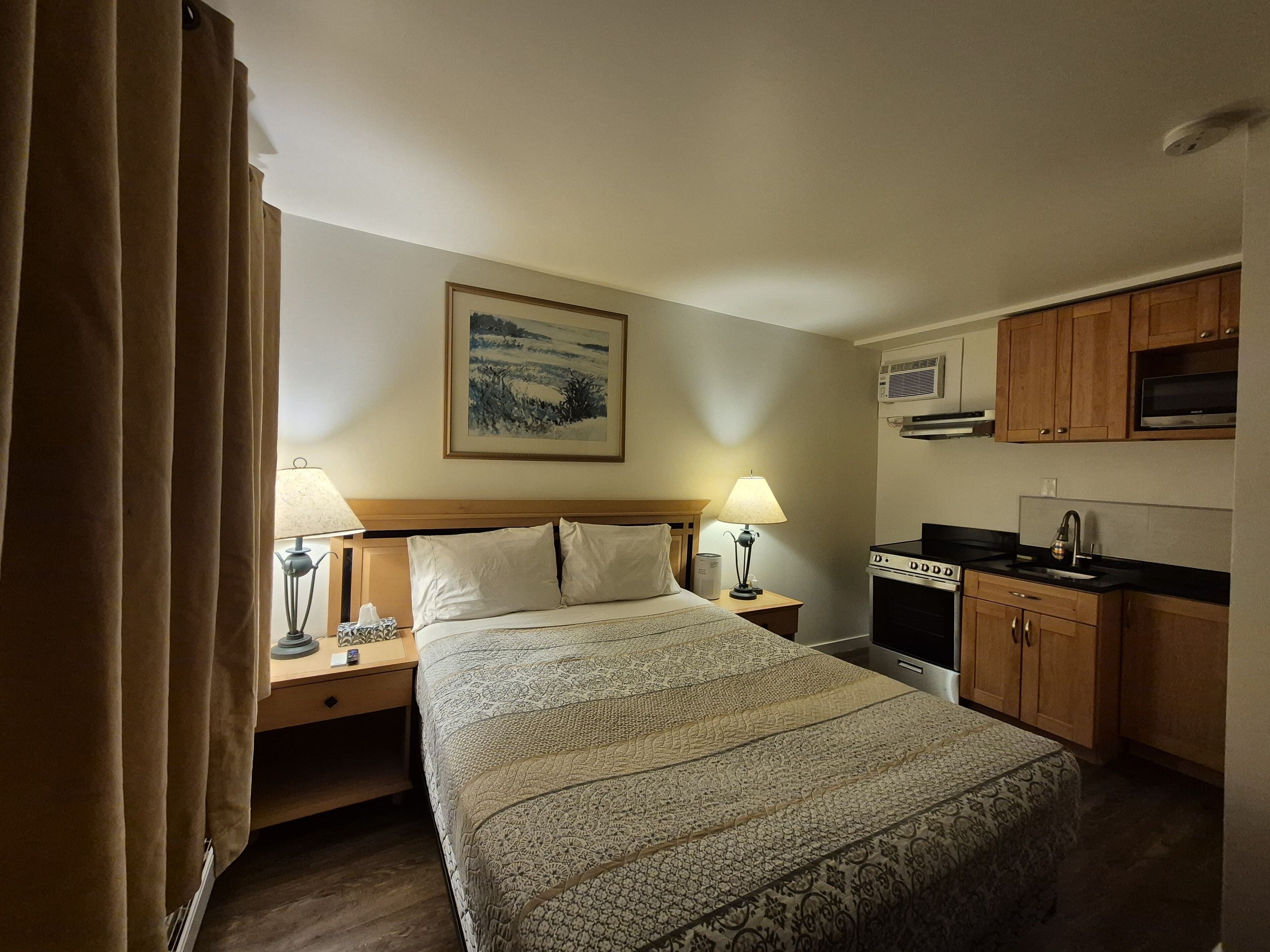 Une chambre d’hôtel comprenant un lit, deux tables de chevet avec des lampes, un tableau au mur, une kitchenette avec une cuisinière et des placards en bois.